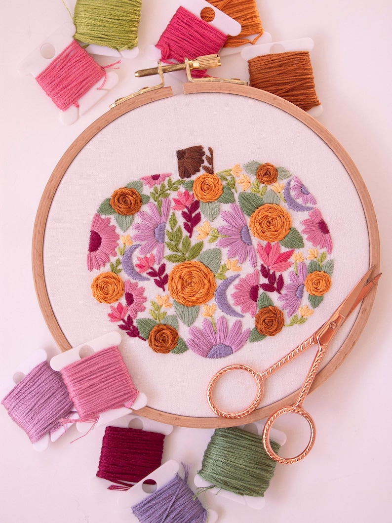 Op de afbeelding: Een borduurring met een bloemenpompoenontwerp in tinten oranje, roze, paars en groen. Spoelen borduurgaren in diverse kleuren en een gouden schaar zijn rond de ring geplaatst.