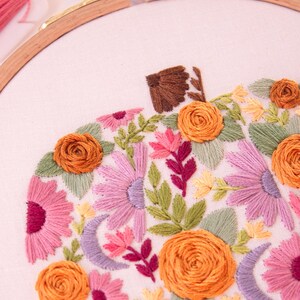 Op de afbeelding: Geborduurd pompoenontwerp op witte stof, in een houten borduurring. De pompoen heeft oranje, roze en paarse bloemen, groene bladeren en een bruine steel. Borduurgaren in verschillende kleuren is zichtbaar.