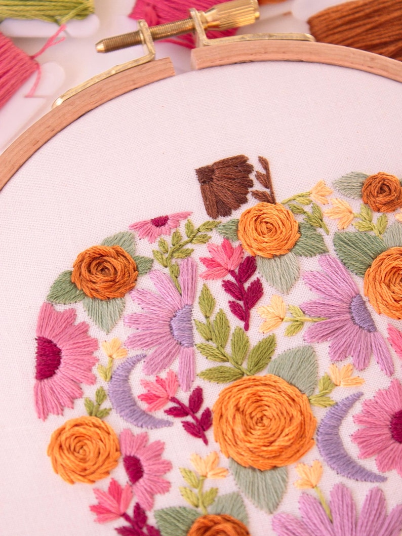 Op de afbeelding: Geborduurd pompoenontwerp op witte stof, in een houten borduurring. De pompoen heeft oranje, roze en paarse bloemen, groene bladeren en een bruine steel. Borduurgaren in verschillende kleuren is zichtbaar.