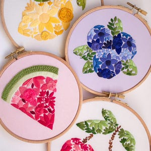 May include: Four embroidery hoops with floral and fruit designs. One features a yellow flower arrangement, another blue flowers, a watermelon slice, and a pair of cherries. Each design is set against a light-colored fabric within a wooden hoop.