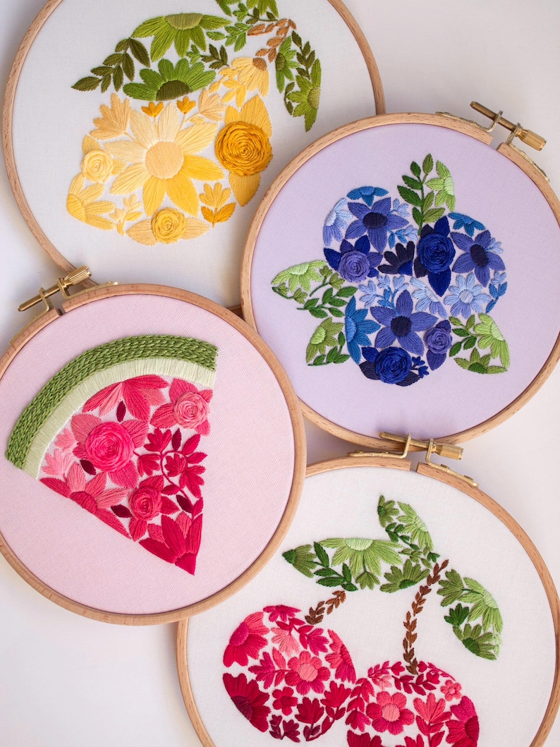 May include: Four embroidery hoops with floral and fruit designs. One features a yellow flower arrangement, another blue flowers, a watermelon slice, and a pair of cherries. Each design is set against a light-colored fabric within a wooden hoop.