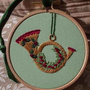 May include: Embroidered Christmas ornament featuring a French horn design. The horn is stitched with gold thread and adorned with green leaves and red berries. The ornament is set on a light green fabric within a wooden hoop, with a green velvet ribbon for hanging.