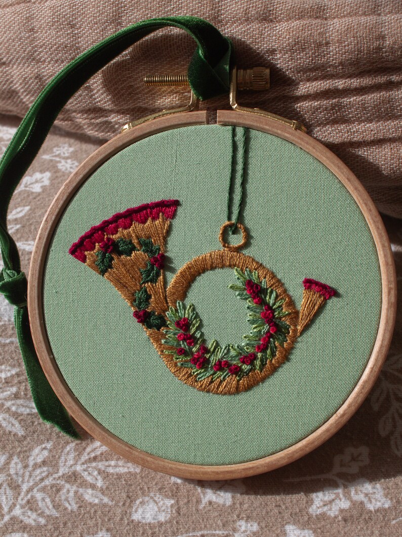 May include: Embroidered Christmas ornament featuring a French horn design. The horn is stitched with gold thread and adorned with green leaves and red berries. The ornament is set on a light green fabric within a wooden hoop, with a green velvet ribbon for hanging.