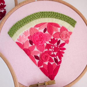May include: A pink fabric embroidery hoop featuring a floral design. The design includes a green arch at the top and a fan-shaped arrangement of pink and red flowers. The embroidery is set in a wooden hoop.