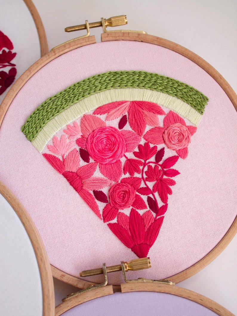 May include: A pink fabric embroidery hoop featuring a floral design. The design includes a green arch at the top and a fan-shaped arrangement of pink and red flowers. The embroidery is set in a wooden hoop.