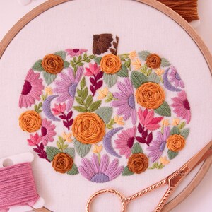 Op de afbeelding: Een borduurring met een pompoenontwerp, gevuld met oranje, roze en paarse bloemen, groene bladeren en halve manen. Roze en bruine garenklosjes en een gouden schaar zijn ook zichtbaar. De ring heeft een diameter van ongeveer 20 cm.