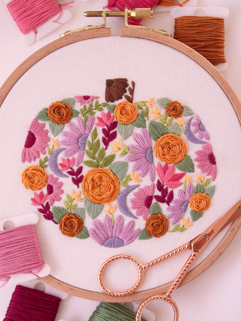 Op de afbeelding: Een borduurring met een pompoenontwerp, gevuld met oranje, roze en paarse bloemen, groene bladeren en halve manen. Roze en bruine garenklosjes en een gouden schaar zijn ook zichtbaar. De ring heeft een diameter van ongeveer 20 cm.