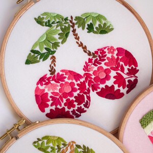 May include: Embroidered cherry design on white fabric stretched within a wooden hoop. The cherries are filled with red and pink floral patterns, with green leaves and brown stems. The embroidery is a craft project.