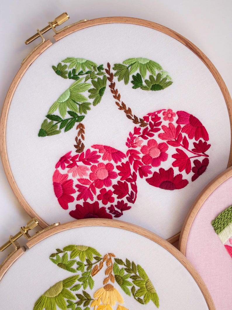 May include: Embroidered cherry design on white fabric stretched within a wooden hoop. The cherries are filled with red and pink floral patterns, with green leaves and brown stems. The embroidery is a craft project.