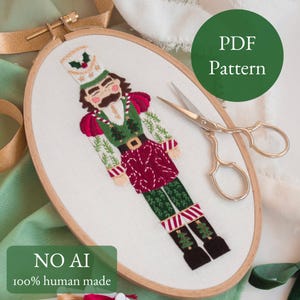 May include: An embroidered nutcracker design in an oval wooden hoop. The nutcracker wears a green and red uniform with a gold belt. The text "PDF Pattern" is in a green circle. The text "NO AI 100% human made" is in a green rectangle. Gold scissors are next to the hoop.