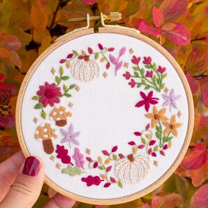May include: A circular wooden embroidery hoop containing white fabric with a hand-stitched wreath design. The wreath features two cream-colored pumpkins, two brown mushrooms, and various autumn-themed floral elements in shades of red, pink, and orange.