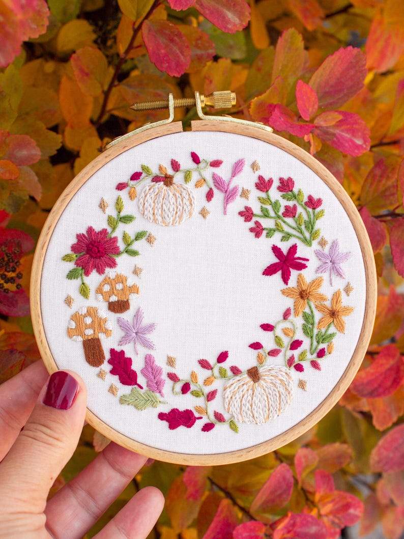 May include: A circular wooden embroidery hoop containing white fabric with a hand-stitched wreath design. The wreath features two cream-colored pumpkins, two brown mushrooms, and various autumn-themed floral elements in shades of red, pink, and orange.