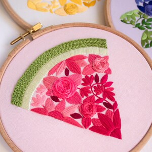 May include: Embroidered watermelon slice in a wooden hoop. The watermelon features pink and red flowers with a green rind. Other hoops with yellow and blue floral designs are visible in the background. The embroidery is on a light pink fabric.