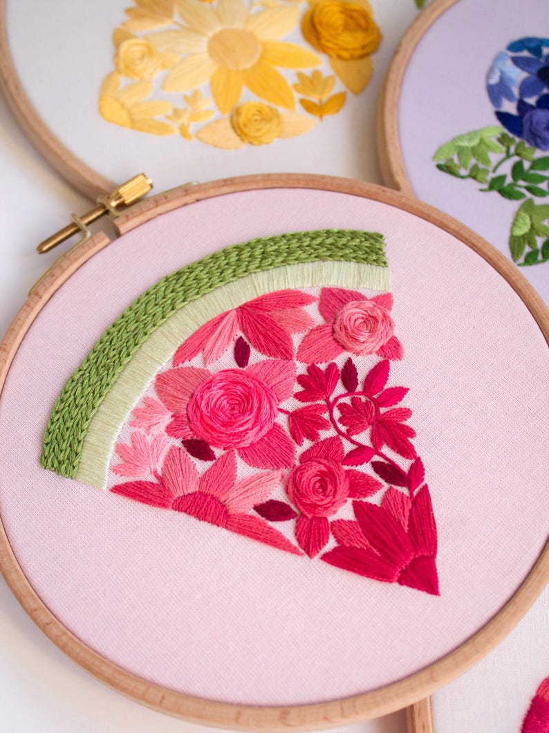 May include: Embroidered watermelon slice in a wooden hoop. The watermelon features pink and red flowers with a green rind. Other hoops with yellow and blue floral designs are visible in the background. The embroidery is on a light pink fabric.