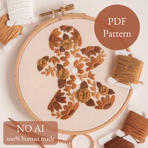 Puede incluir: Un bastidor de bordado con un diseño de hombre de jengibre, con bordados florales marrones y beige. El texto "PDF Pattern" está en un círculo marrón. El texto "NO AI 100% human made" está en la parte inferior. También se ven carretes de hilo y tijeras.
