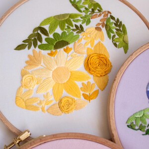May include: Embroidered floral design on white fabric stretched within a wooden hoop. The design features yellow and green flowers and leaves. The hoop is secured with a brass screw. Other hoops with floral designs are visible.