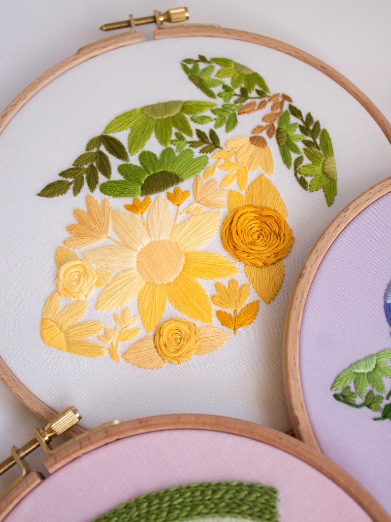 May include: Embroidered floral design on white fabric stretched within a wooden hoop. The design features yellow and green flowers and leaves. The hoop is secured with a brass screw. Other hoops with floral designs are visible.