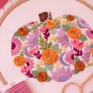Op de afbeelding: Een geborduurde pompoen in een houten ring. De pompoen is gevuld met kleurrijke bloemen, bladeren en halve manen in roze, oranje, paarse en groene tinten. Borduurgaren en schaar zijn zichtbaar.