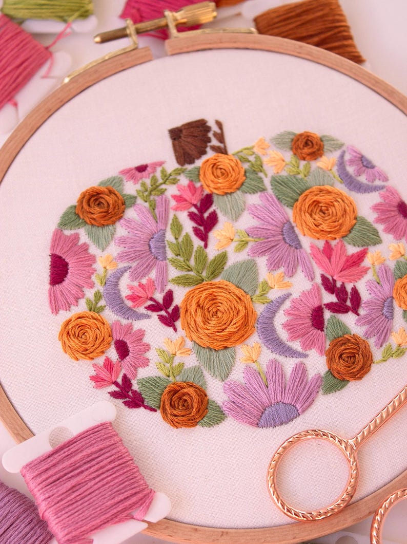 Op de afbeelding: Een geborduurde pompoen in een houten ring. De pompoen is gevuld met kleurrijke bloemen, bladeren en halve manen in roze, oranje, paarse en groene tinten. Borduurgaren en schaar zijn zichtbaar.