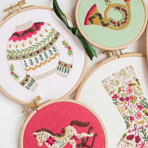 May include: Embroidered Christmas ornaments in wooden hoops. Designs include a sweater, a French horn, a rocking horse, and a stocking. The ornaments feature red, green, and gold embroidery on white, red, and green backgrounds. A green ribbon is attached to one ornament.