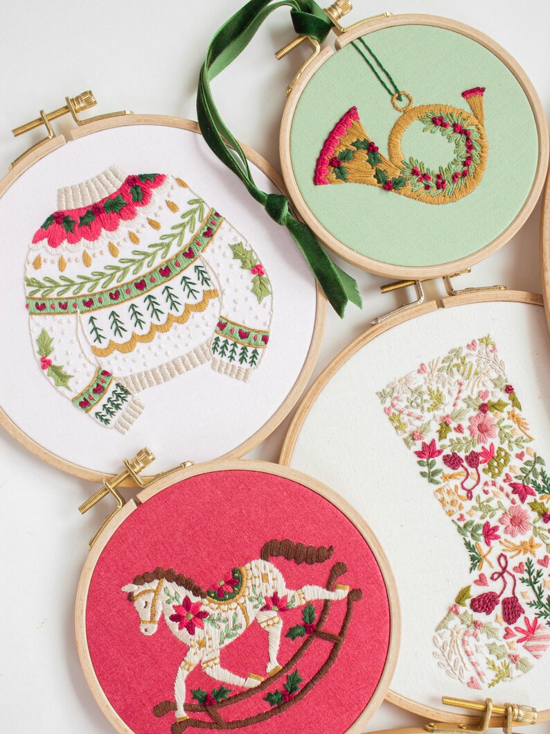 May include: Embroidered Christmas ornaments in wooden hoops. Designs include a sweater, a French horn, a rocking horse, and a stocking. The ornaments feature red, green, and gold embroidery on white, red, and green backgrounds. A green ribbon is attached to one ornament.