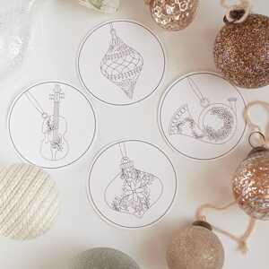 May include: Overhead shot of Christmas ornaments and decorative coasters. The coasters feature line drawings of ornaments, a violin, and a French horn. The ornaments are in various colors, including gold, silver, and green. The background is white.