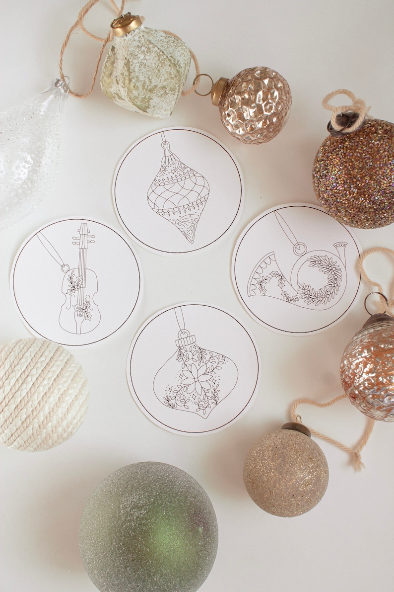 May include: Overhead shot of Christmas ornaments and decorative coasters. The coasters feature line drawings of ornaments, a violin, and a French horn. The ornaments are in various colors, including gold, silver, and green. The background is white.