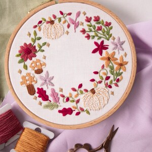 May include: A circular wooden embroidery hoop containing white fabric with a hand-stitched wreath design. The embroidery features two cream-colored pumpkins, various autumn-toned flowers, green leaves, and two spotted mushrooms in shades of brown.