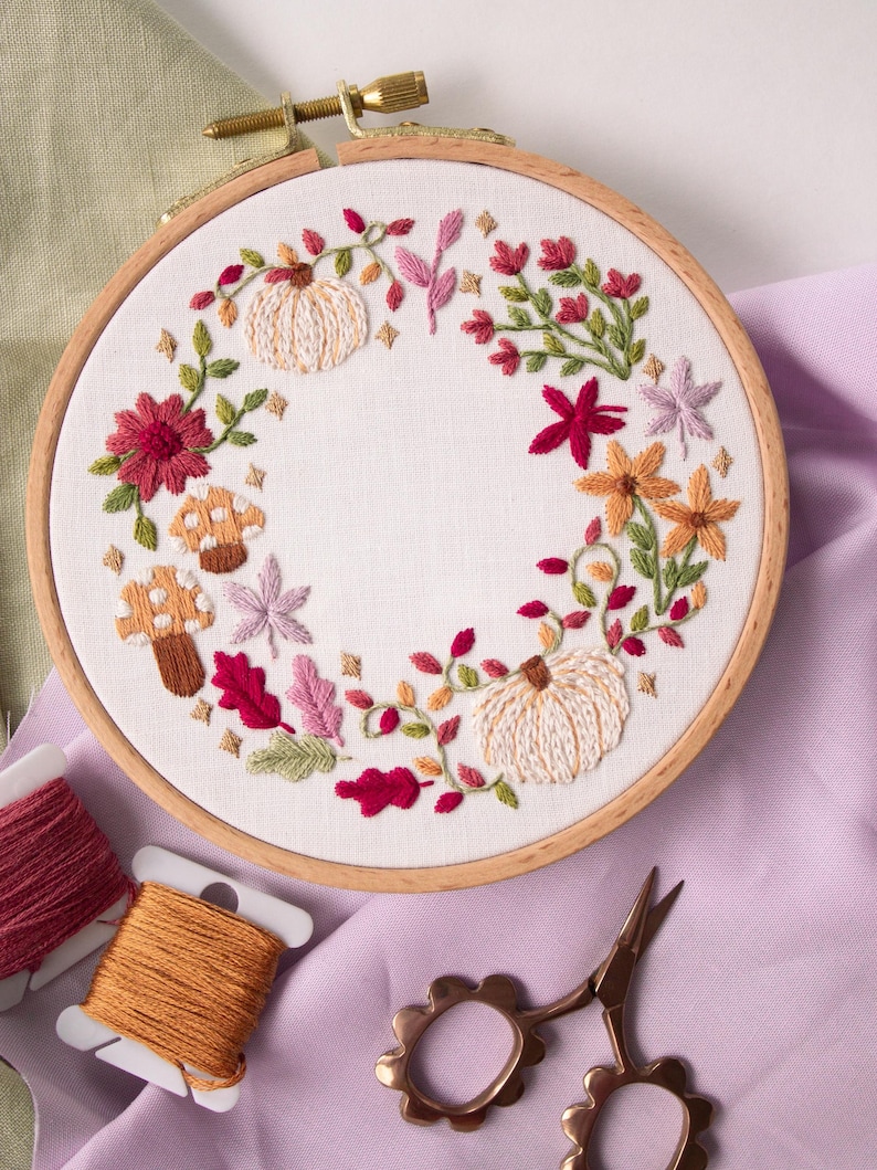 May include: A circular wooden embroidery hoop containing white fabric with a hand-stitched wreath design. The embroidery features two cream-colored pumpkins, various autumn-toned flowers, green leaves, and two spotted mushrooms in shades of brown.