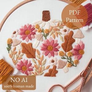 May include: An embroidery hoop containing white fabric with a stitched pumpkin design composed of pink flowers, brown oak leaves, small mushrooms, and yellow buds. The image includes text reading PDF Pattern and NO AI 100% human made.