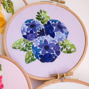 May include: Embroidered floral hoop art featuring a central arrangement of blue flowers and green leaves on a lavender fabric. The embroidery is set in a wooden hoop. Other hoops with floral designs are visible.