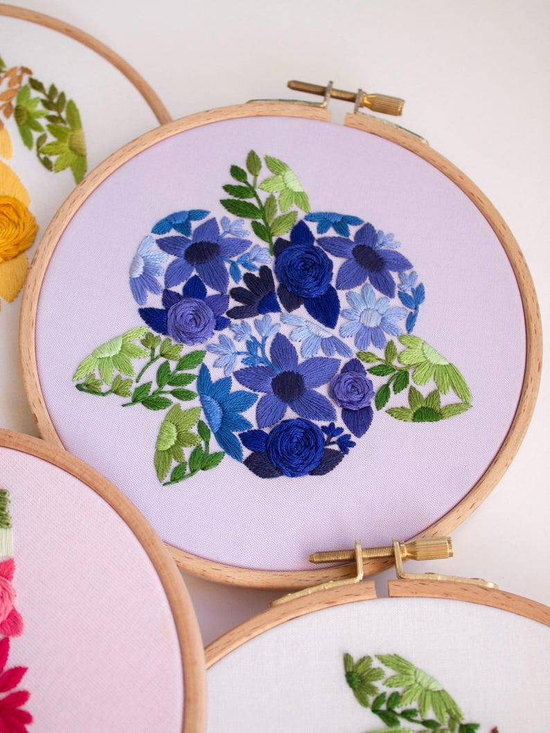 May include: Embroidered floral hoop art featuring a central arrangement of blue flowers and green leaves on a lavender fabric. The embroidery is set in a wooden hoop. Other hoops with floral designs are visible.