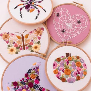 Op de afbeelding: Een verzameling van vijf borduurringen met Halloween- en herfstthema's. De ontwerpen omvatten een spin, een spinnenwebhart, een vlinder, een heksenhoed en een pompoen, allemaal met bloemen- en blad details.