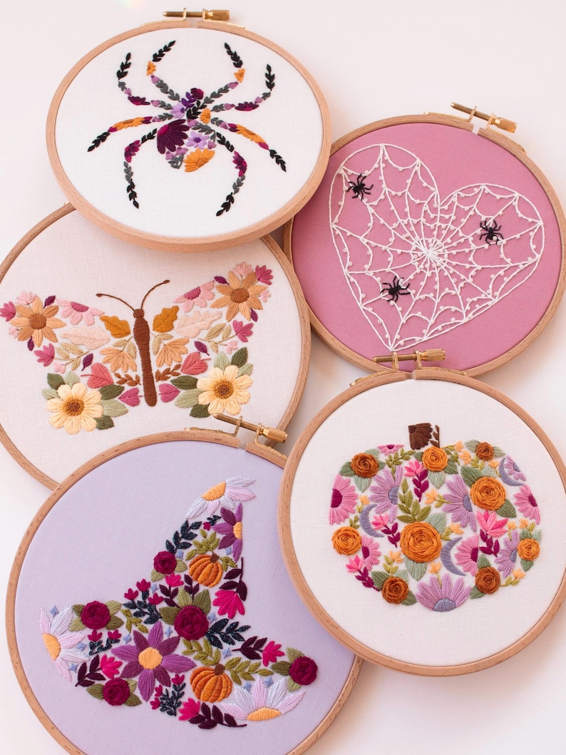 Op de afbeelding: Een verzameling van vijf borduurringen met Halloween- en herfstthema's. De ontwerpen omvatten een spin, een spinnenwebhart, een vlinder, een heksenhoed en een pompoen, allemaal met bloemen- en blad details.