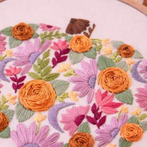 Op de afbeelding: Geborduurd pompoenontwerp met oranje rozen, roze en paarse bloemen en groene bladeren. De borduursels zijn geplaatst in een houten hoepel, met een herfstthema. Het ontwerp bevat een bruine stengel en halve maan vormen.