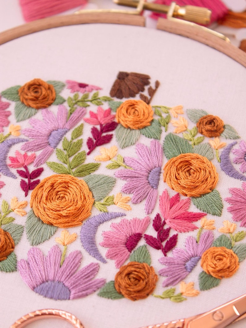 Op de afbeelding: Geborduurd pompoenontwerp met oranje rozen, roze en paarse bloemen en groene bladeren. De borduursels zijn geplaatst in een houten hoepel, met een herfstthema. Het ontwerp bevat een bruine stengel en halve maan vormen.