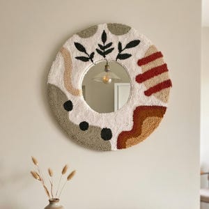 Puede incluir: Un espejo redondo decorativo con un borde texturizado blanquecino con formas abstractas en negro, marrón y rojo. Cuelga en una pared neutra sobre una mesa de madera con un jarrón y libros.