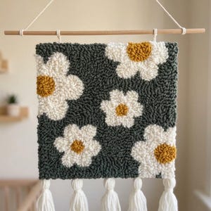 Op de afbeelding: Een vierkante, handgemaakte wandhanger met een donkergroene achtergrond en witte madeliefbloemen met gele centra. Opgehangen aan een houten stok met witte kwastjes. Een decoratief item voor een kinderkamer, ongeveer 30 cm x 30 cm.