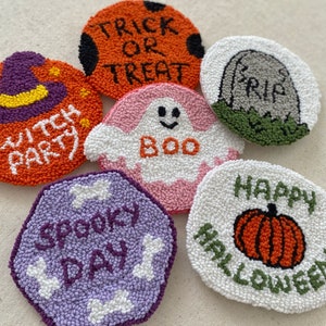 May include: Six handmade Halloween-themed punch needle coasters. The coasters are round and feature various designs, including a ghost, a pumpkin, a witch's hat, a tombstone, and a bone. The coasters are made with colorful yarn and have a variety of textures.