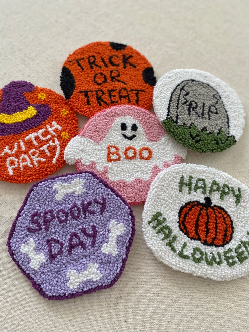 May include: Six handmade Halloween-themed punch needle coasters. The coasters are round and feature various designs, including a ghost, a pumpkin, a witch's hat, a tombstone, and a bone. The coasters are made with colorful yarn and have a variety of textures.