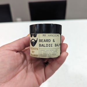 May include: A small, round jar with a black lid and a label that reads "Mr. Handsome Beard & Baldie Balm". The label is beige with black text and a cartoon image of a bearded man wearing sunglasses. The text on the label reads "Scents by Sharif Organic Shea Butter, Jojoba Oil, Vitamin E Oil, Coconut Oil, Babassu Oil, Candelilla Wax, Essential & Premium Fragrance Oils."