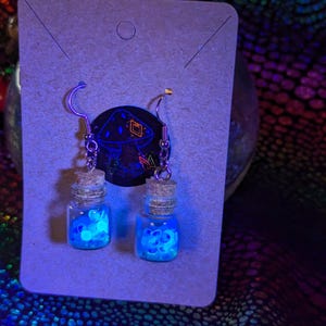 May include: Handmade earrings with miniature glass bottles and cork stoppers. The bottles are filled with glowing blue shapes. The earrings hang from silver hooks and are displayed on a neutral card with a mushroom graphic.