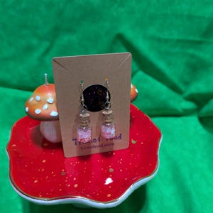 May include: Dangle earrings with small glass bottles filled with pink glitter and cork stoppers. The earrings hang from silver hooks and are displayed on a brown card with a logo and the text "Trinket Toad Trinkettoad.store". The earrings are set on a red mushroom-shaped display.