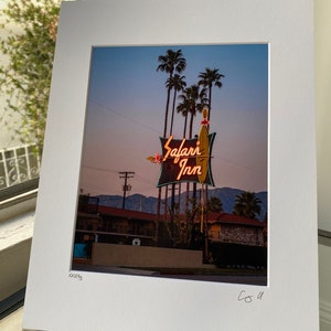 May include: A vintage motel sign with neon lights that reads "Safari Inn" against a twilight sky. Palm trees are in the foreground and a low-rise motel building is in the background.