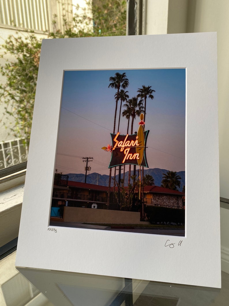 May include: A vintage motel sign with neon lights that reads "Safari Inn" against a twilight sky. Palm trees are in the foreground and a low-rise motel building is in the background.