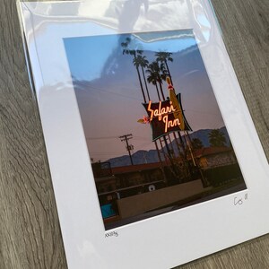 May include: A vintage photograph of a neon sign for the "Safari Inn" motel. The sign is lit up in pink, red, and yellow, and features a palm tree and a surfboard. The photo was taken at dusk, and the sky is a soft blue.