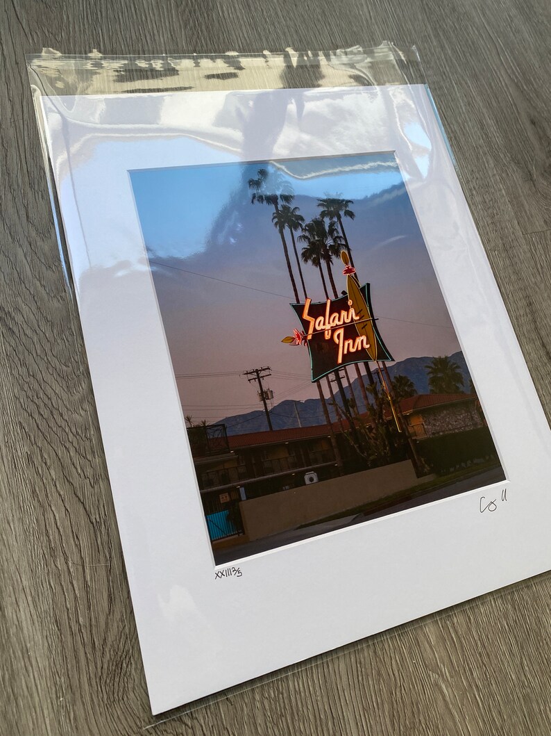 May include: A vintage photograph of a neon sign for the "Safari Inn" motel. The sign is lit up in pink, red, and yellow, and features a palm tree and a surfboard. The photo was taken at dusk, and the sky is a soft blue.