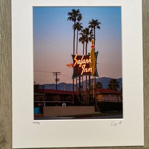 May include: A vintage neon sign for the Safari Inn motel, with palm trees in the background. The sign is lit up in orange and yellow, with the words "Safari Inn" in a stylised font.