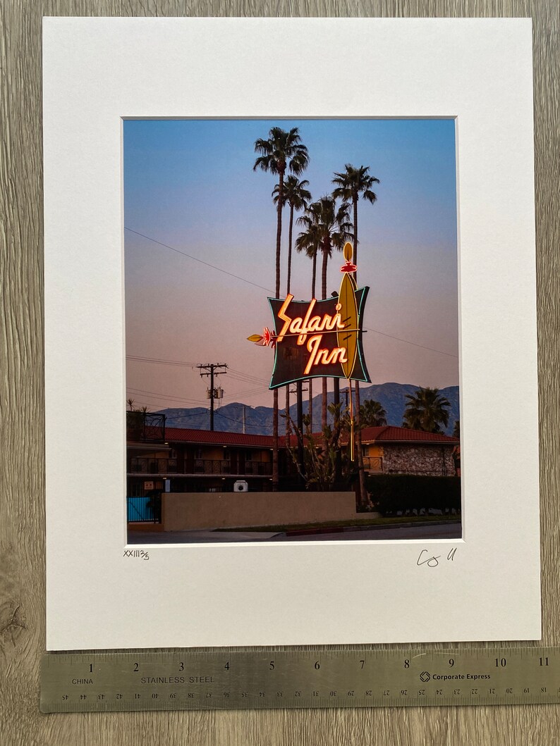 May include: A vintage neon sign for the Safari Inn motel, with palm trees in the background. The sign is lit up in orange and yellow, with the words "Safari Inn" in a stylised font.