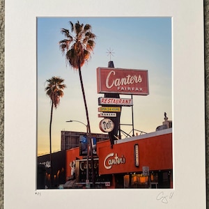 May include: A vintage photograph of Canter's Deli in Los Angeles, California. The restaurant is known for its classic diner atmosphere and delicious food. The image features a bright red building with a large sign that reads "Canter's Fairfax" in white lettering. The sign is illuminated with neon lights. There are palm trees in the foreground and a blue sky in the background.